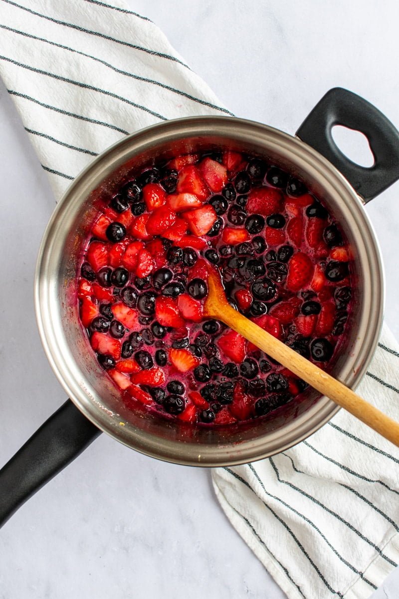 Strawberries and blueberries simmering in a saucepan with a wooden spoon.