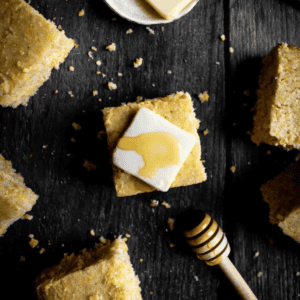 Five squares of cornbread on a dark wood table next to a honey stir stick.