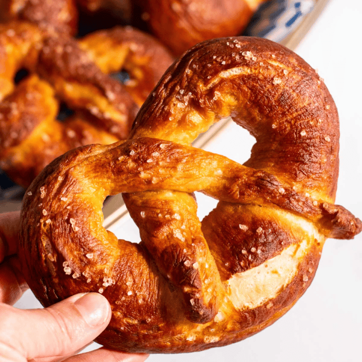 A hand holding a large Bavarian pretzel near a dish of more baked pretzels.