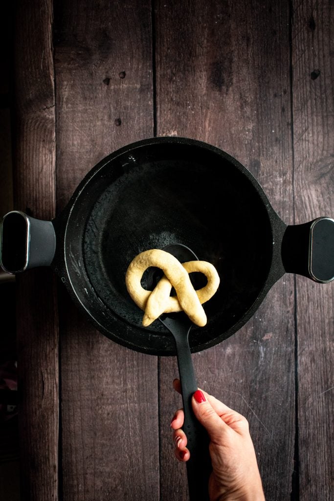 A hand holding an unbaked pretzel over a pot of water.