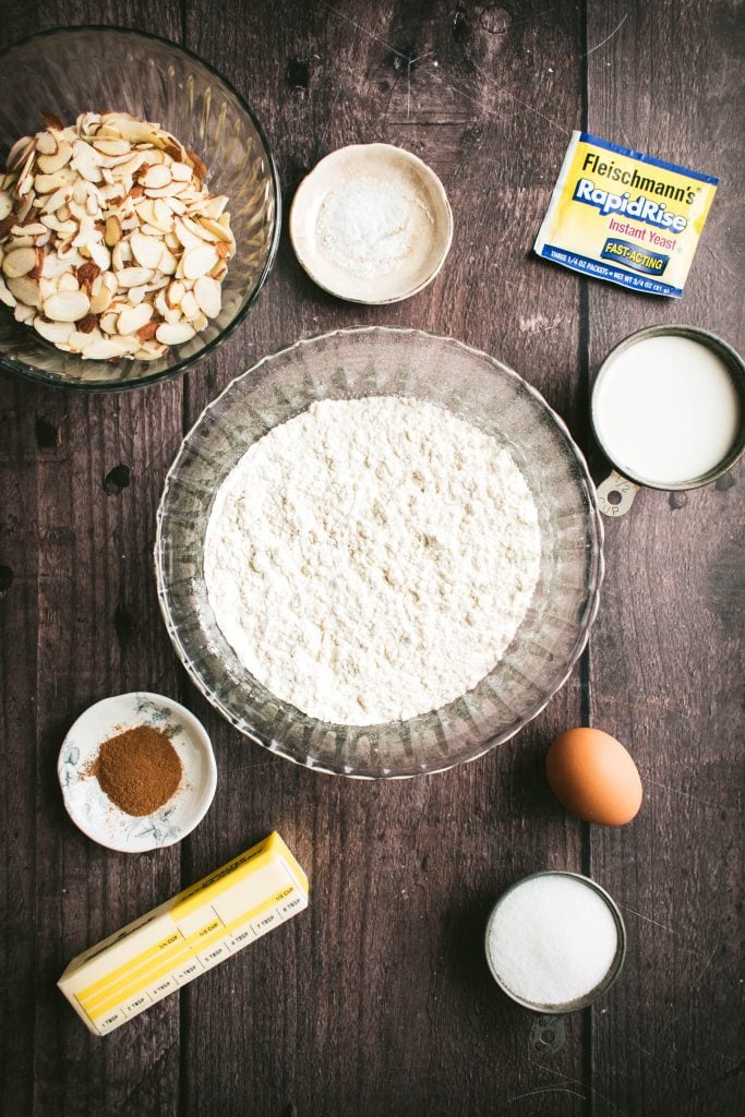 Bowls of flour, cinnamon, milk, salt, and sliced almonds next to butter, eggs, and a packet of yeast on a table.