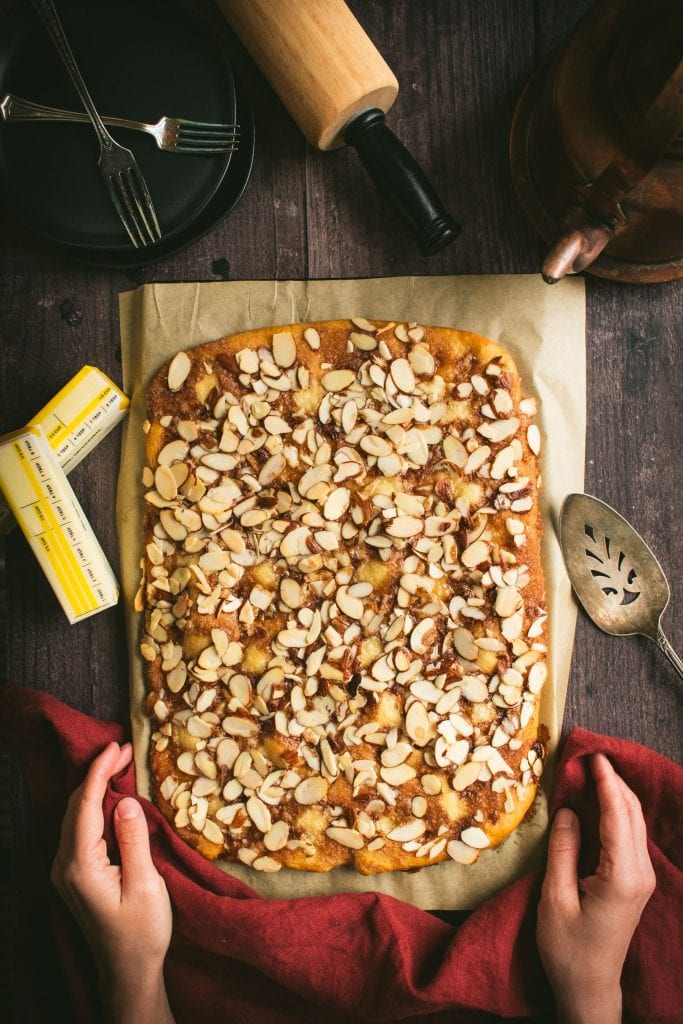 Two hands near a thin, rectangular coffee cake topped with brown sugar and sliced almonds.