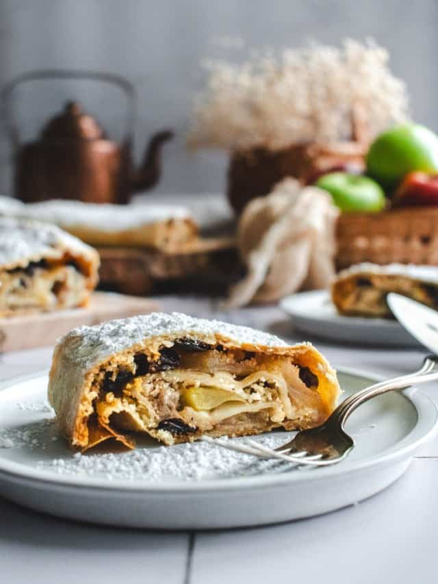 A slice of homemade apple strudel filled with apples and raisins and topped with powdered sugar on a grey plate on a light surface.