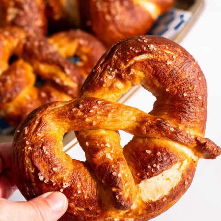 A hand holding a large Bavarian pretzel next to a baking sheet filled with more soft pretzels.