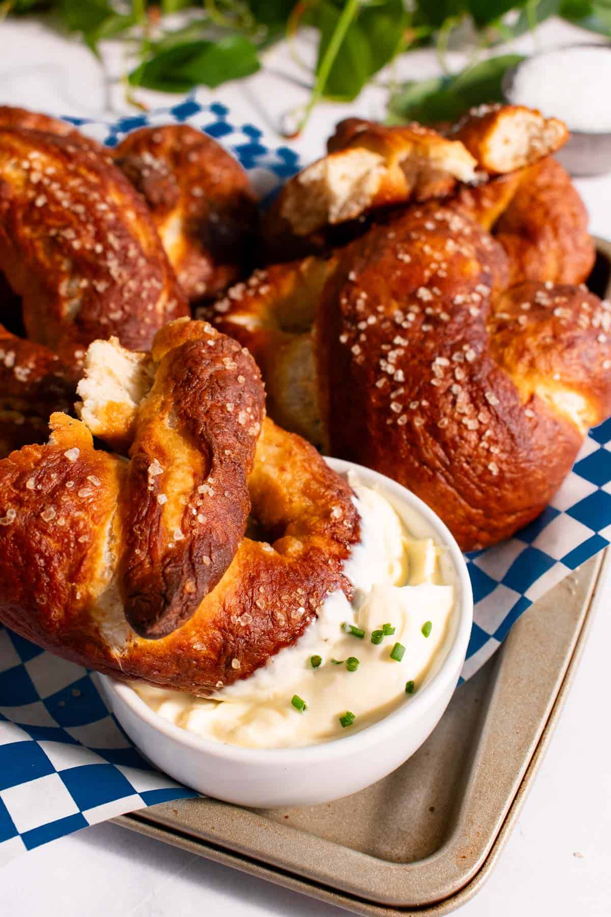 A half-eaten soft pretzel dunked into a bowl of mayo on a baking sheet next to more baked pretzels.
