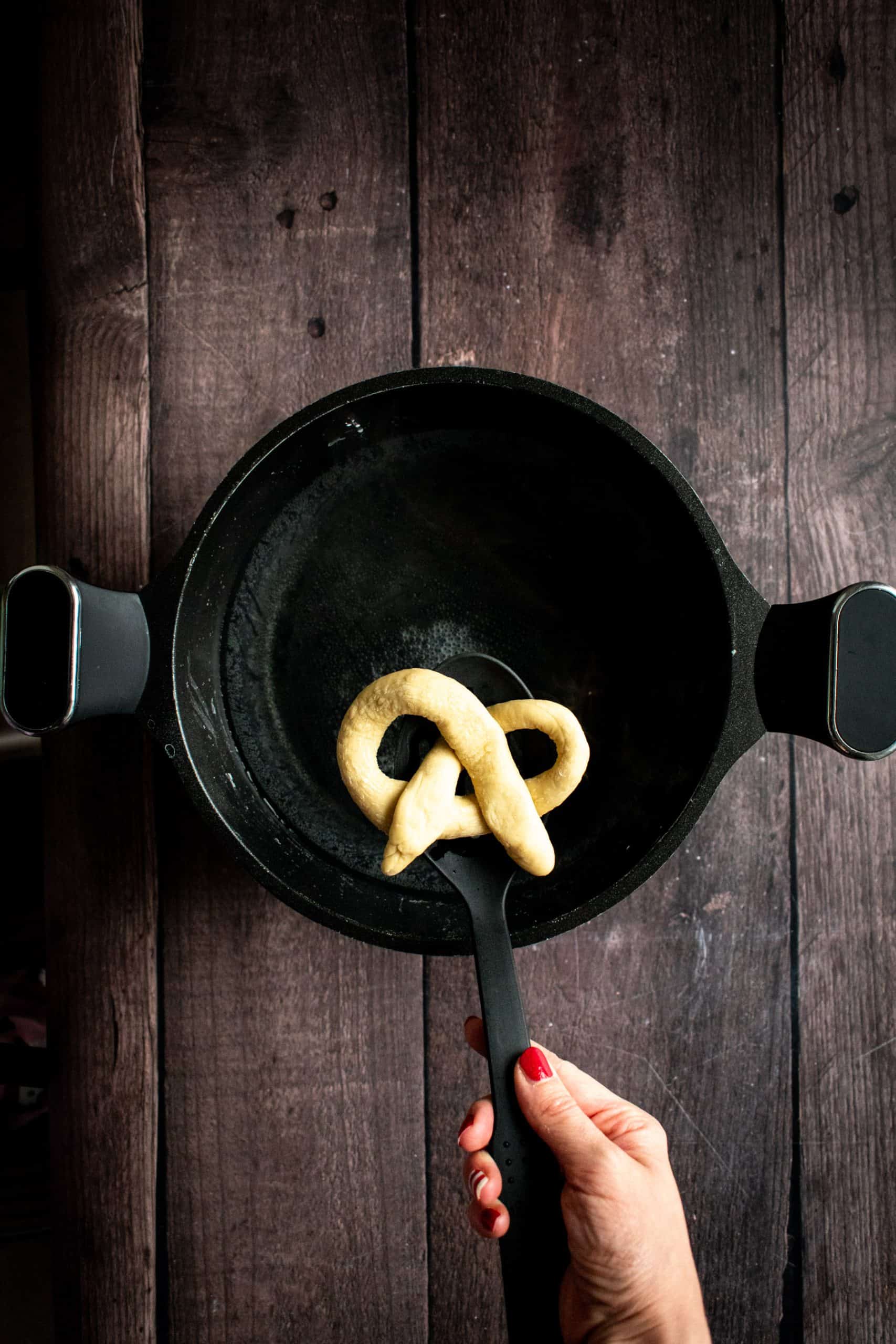 A hand placing an unbaked pretzel in a pot of water with a slotted spoon.