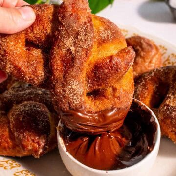 A hand dipping a cinnamon pretzel into a dish of melted chocolate.