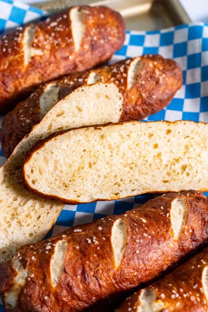 A hot dog bun cut in half to show the soft inside, on a baking sheet next to four other pretzel bread sticks.