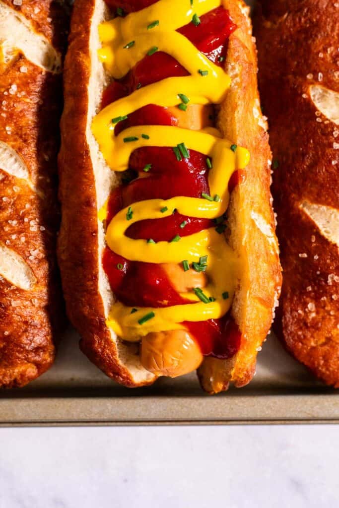 A hot dog topped with mustard and ketchup in a golden brown pretzel hot dog bun between two more buns on a metal tray.
