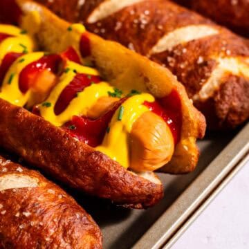 Pretzel hot dog buns on a baking sheet, with one filled with a hot dog topped with mustard and ketchup.