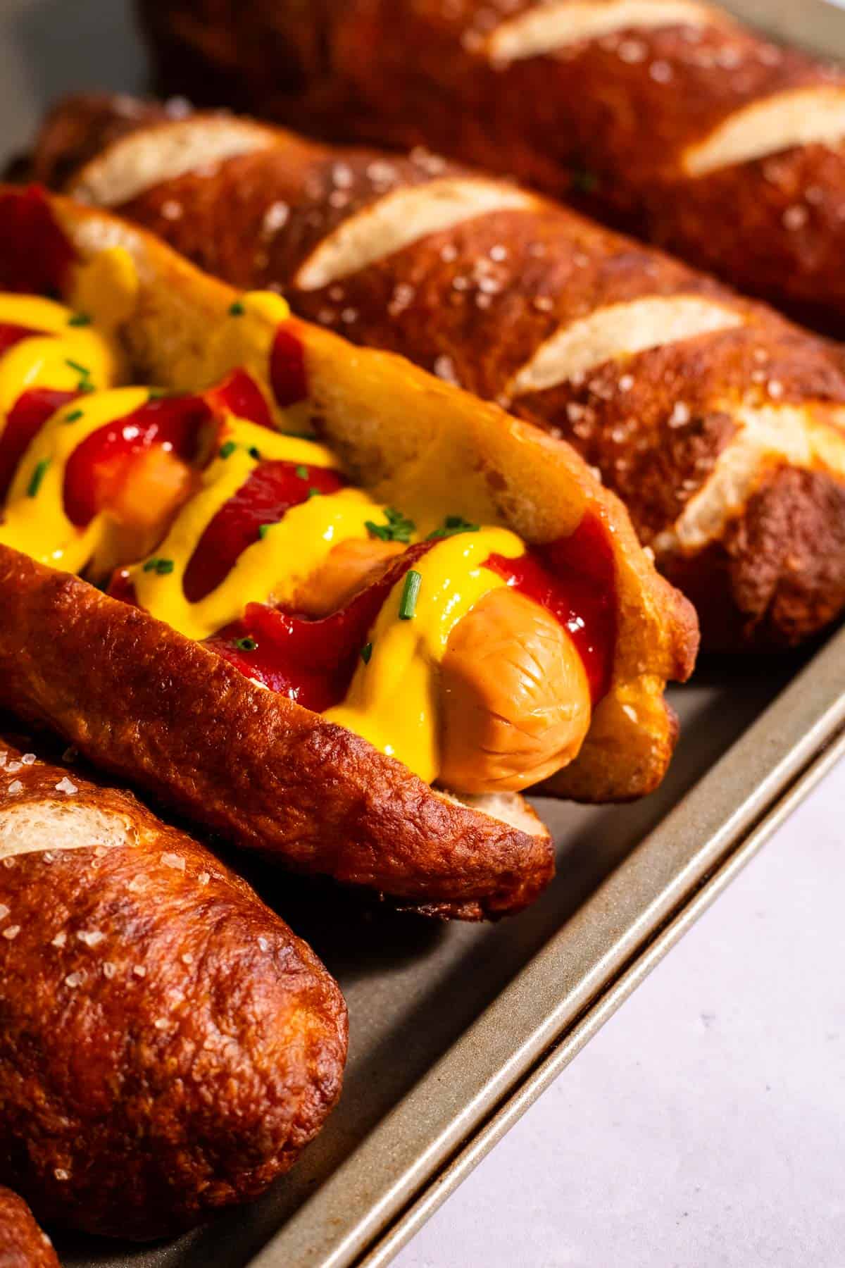 Pretzel hot dog buns on a baking sheet, with one filled with a hot dog topped with mustard and ketchup.