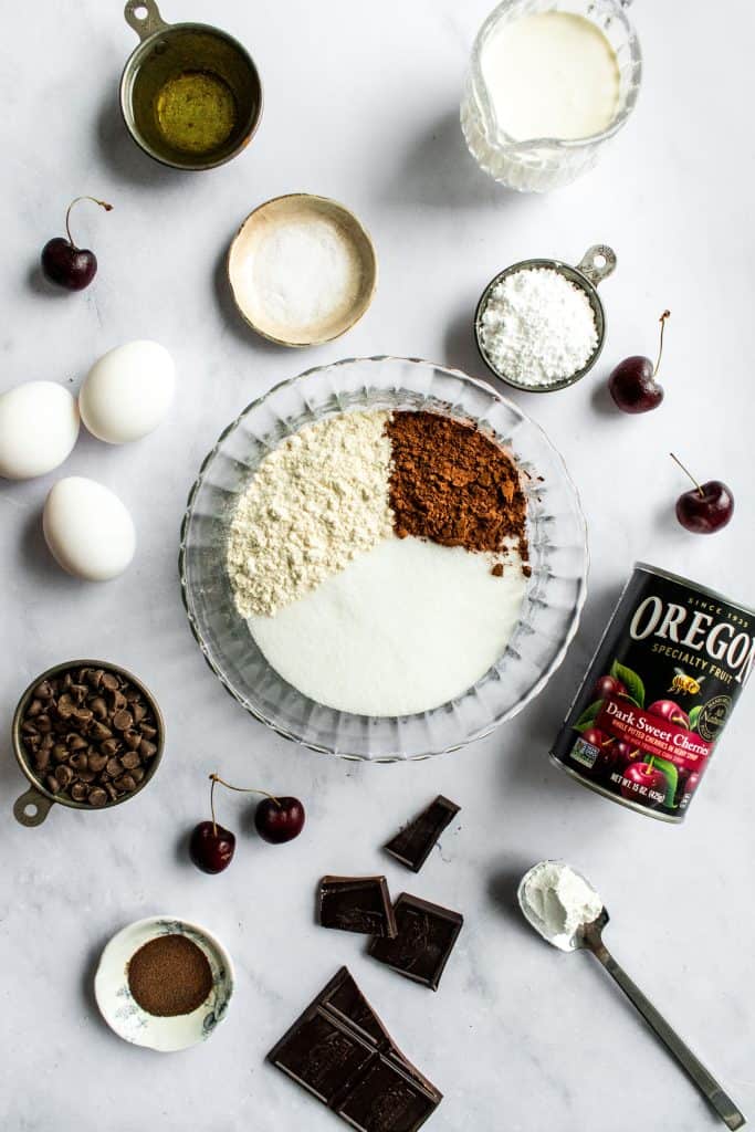 Flour, sugar, and cocoa powder in a glass bowl next to other raw ingredients.