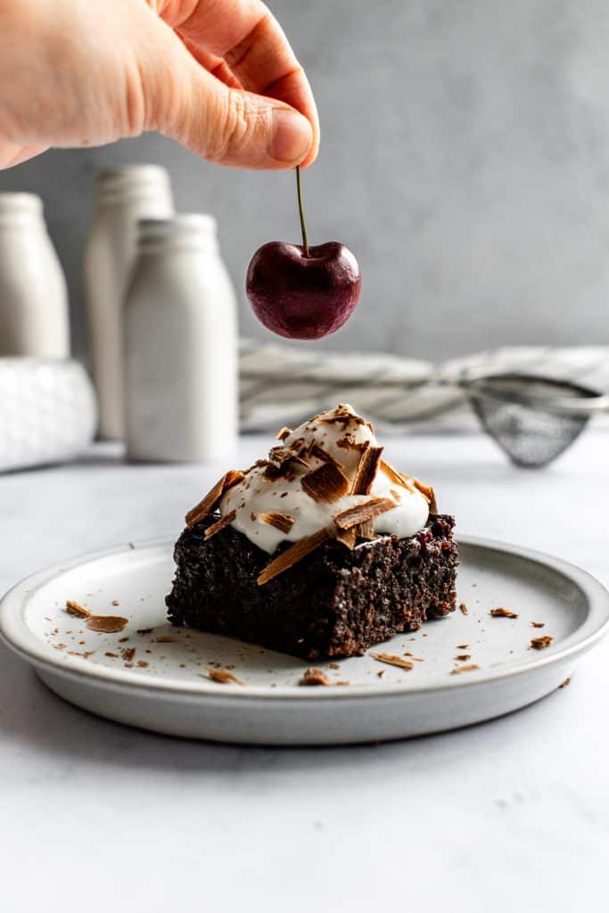 A hand placing a fresh cherry onto a brownie with whipped cream.