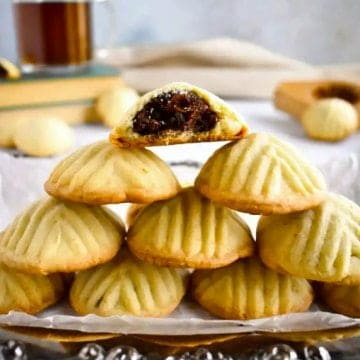 Half a dozen butter cookies piled on a silver plate, with the top one cut in half to show the date filling.