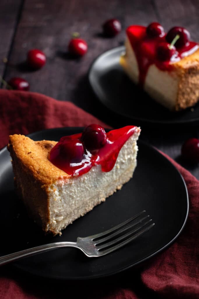 A slice of creamy cheesecake topped with cherry sauce and cherries on a small black plate with a fork.