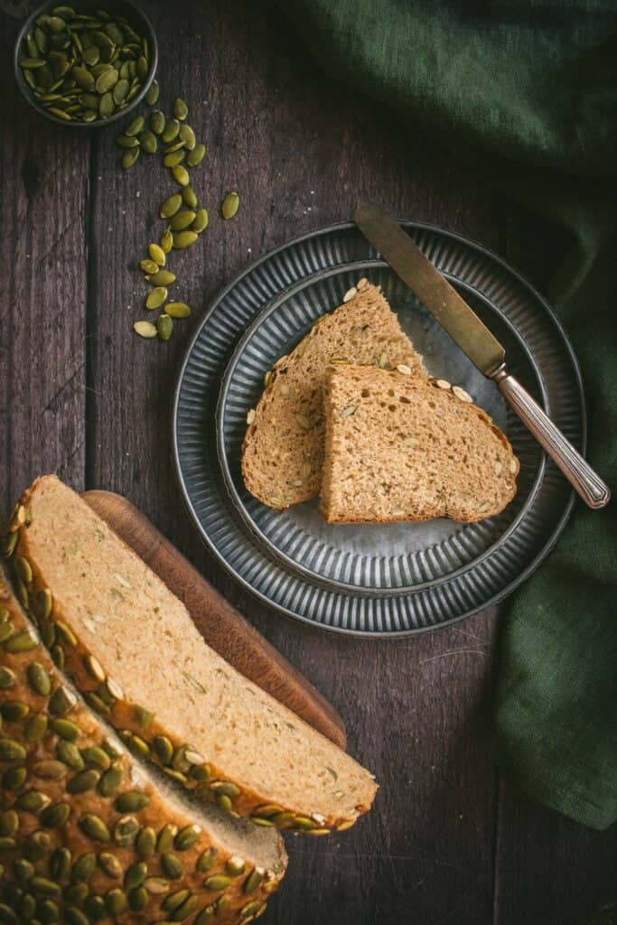 Slices of hearty brown bread on a metal plate next to the rest of the loaf topped with hulled pumpkin seeds.