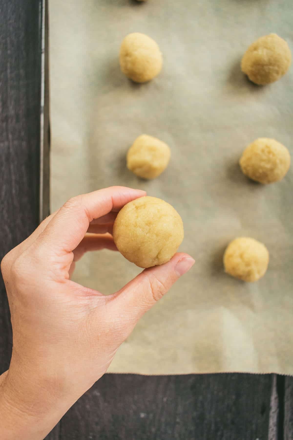 A hand holding a small round ball of light beige cookie dough over a baking sheet.