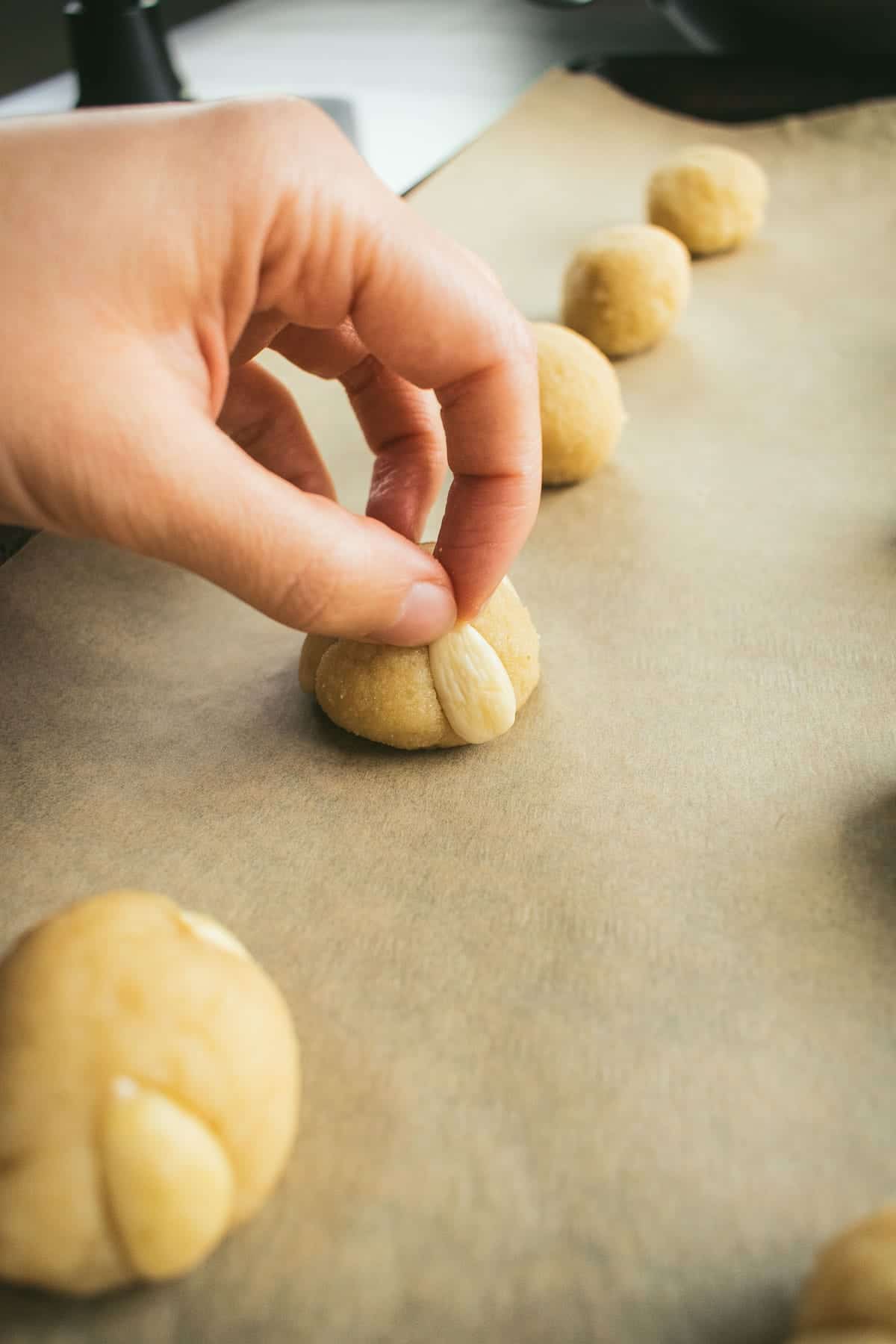 A hand pressing sliced almonds into the sides of cookie dough balls on a baking sheet.