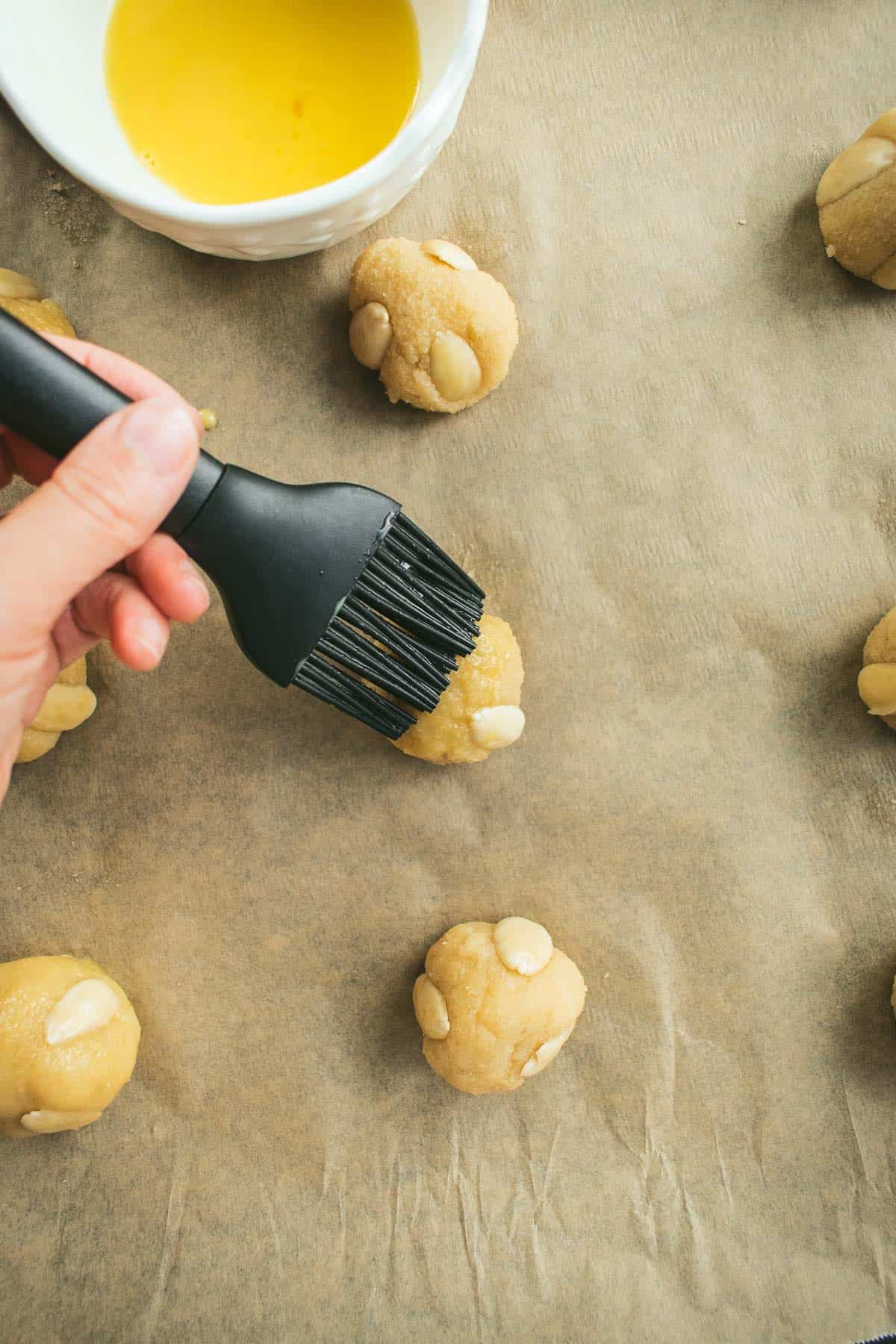 A hand brushing egg wash onto small cookie dough balls.