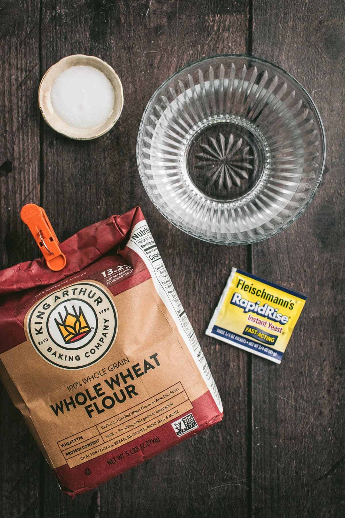A bag of whole wheat flour on a table next to instant yeast and bowls of water and salt.
