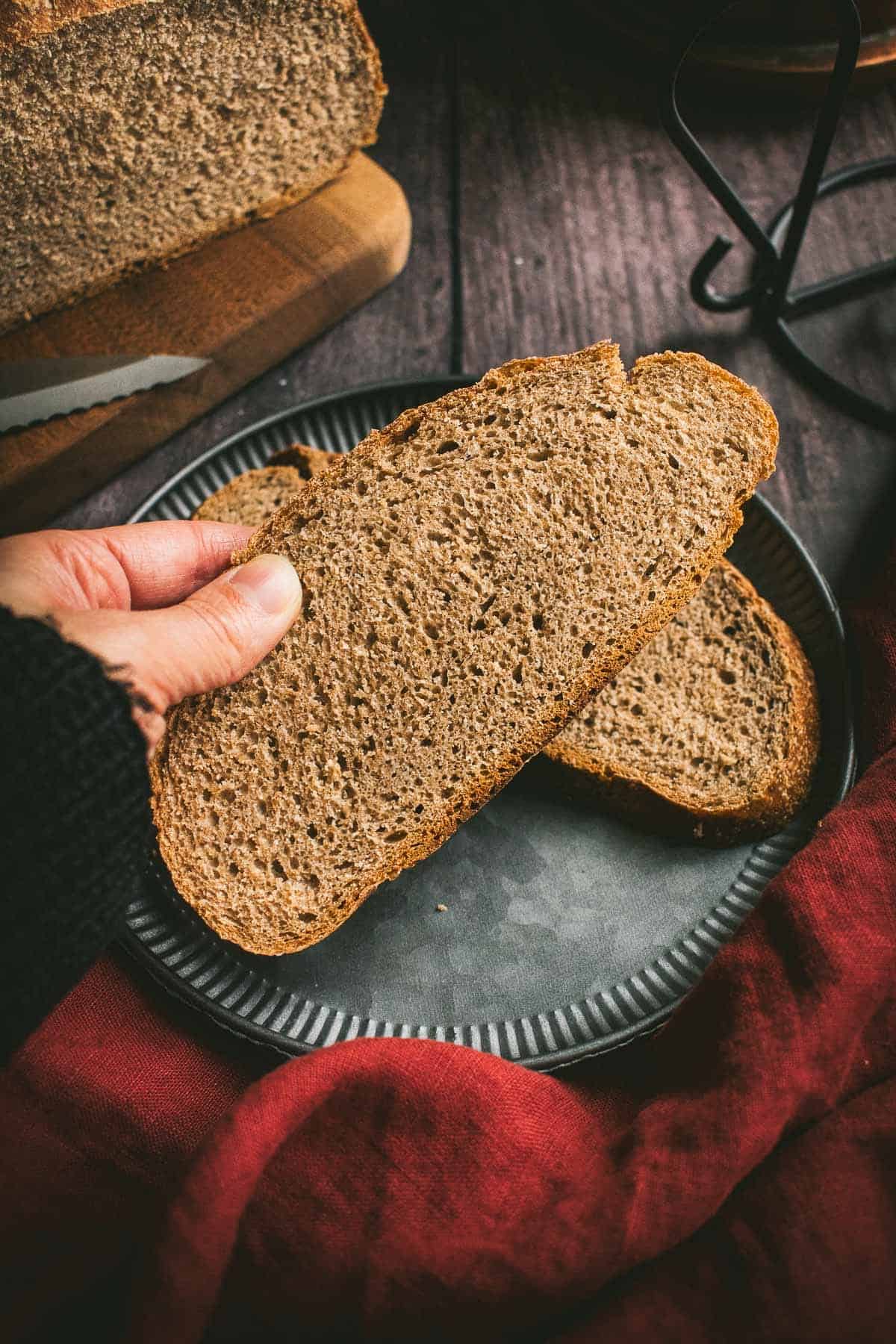 A hand holding a large slice of dark brown bread over a red linen.