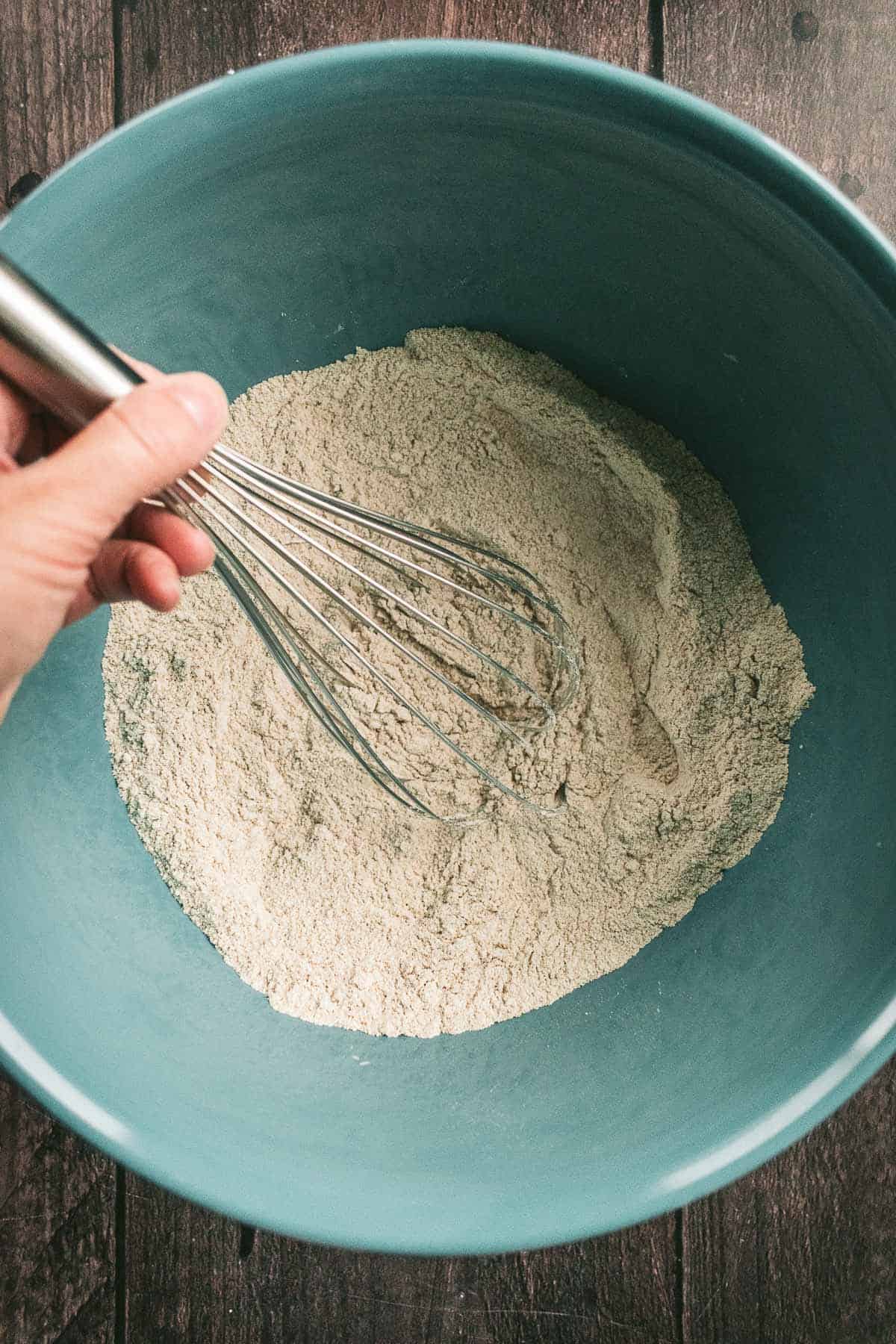 A hand whisking flour, salt, and yeast in a large mixing bowl.
