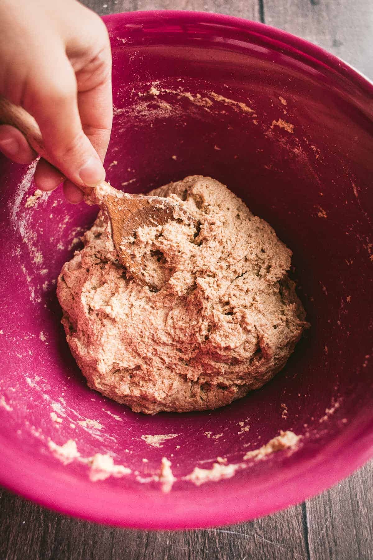 A hand mixing bread dough in a mixing bowl with a wooden spoon.