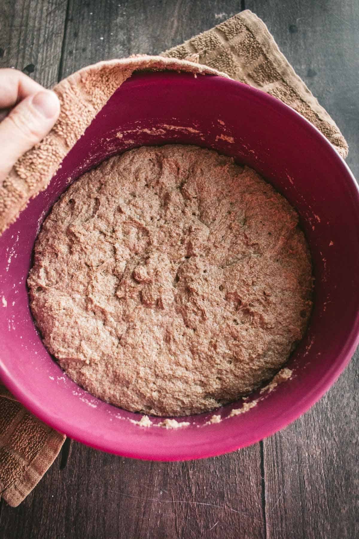 A hand removing a towel from the top of a mixing bowl with risen wheat bread dough.