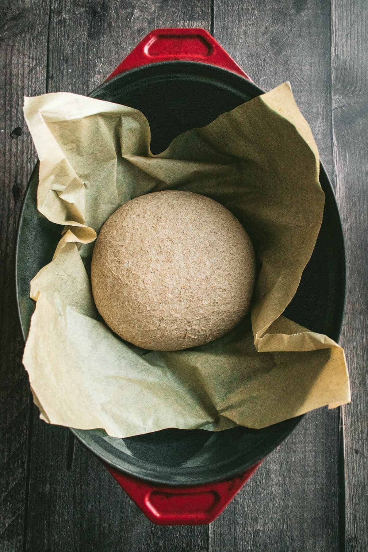 A ball of light beige bread dough on parchment paper in a Dutch oven.