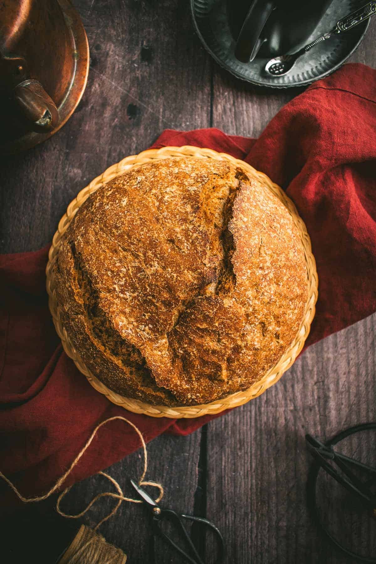 A baked loaf of crusty bread on a wicker trivet on a red linen.