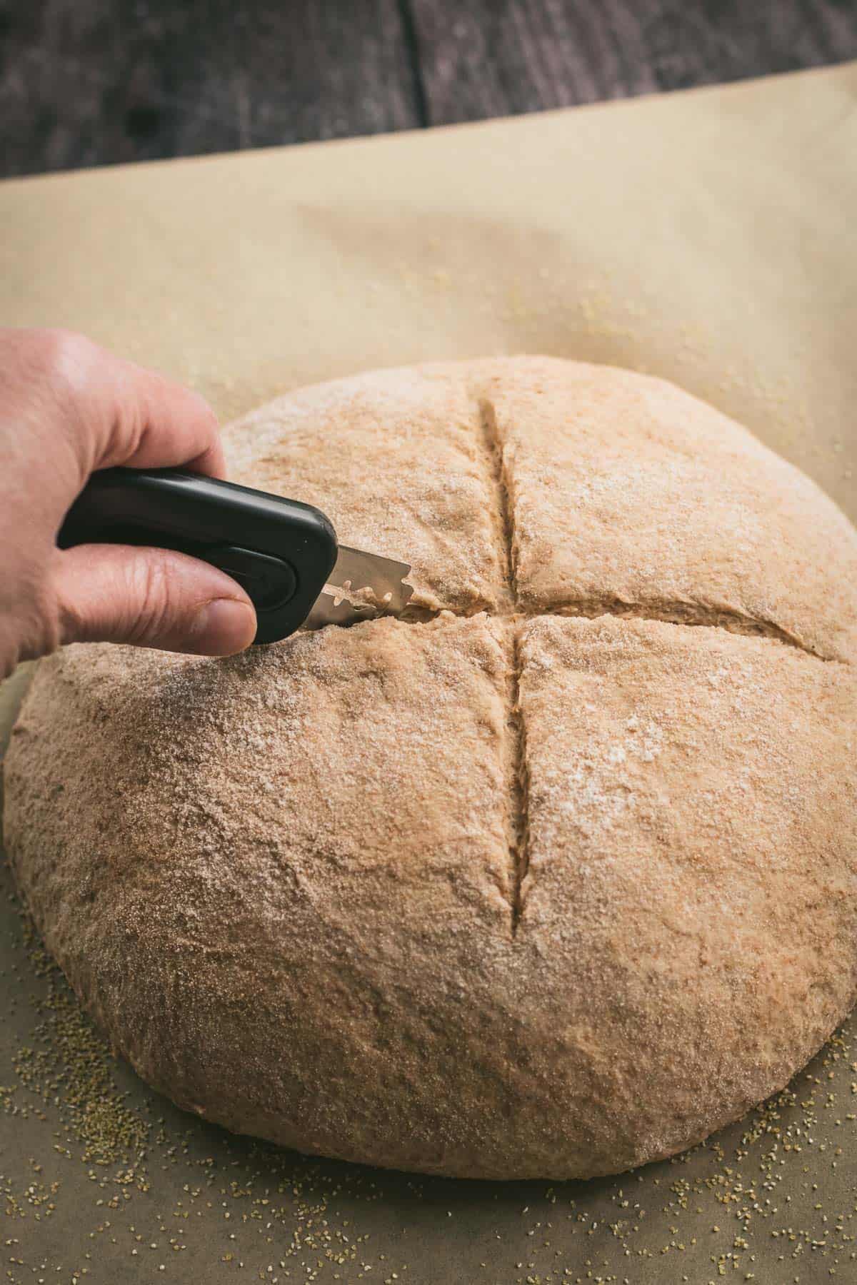 A hand scoring a boule of bread dough on parchment paper with a bread lame.