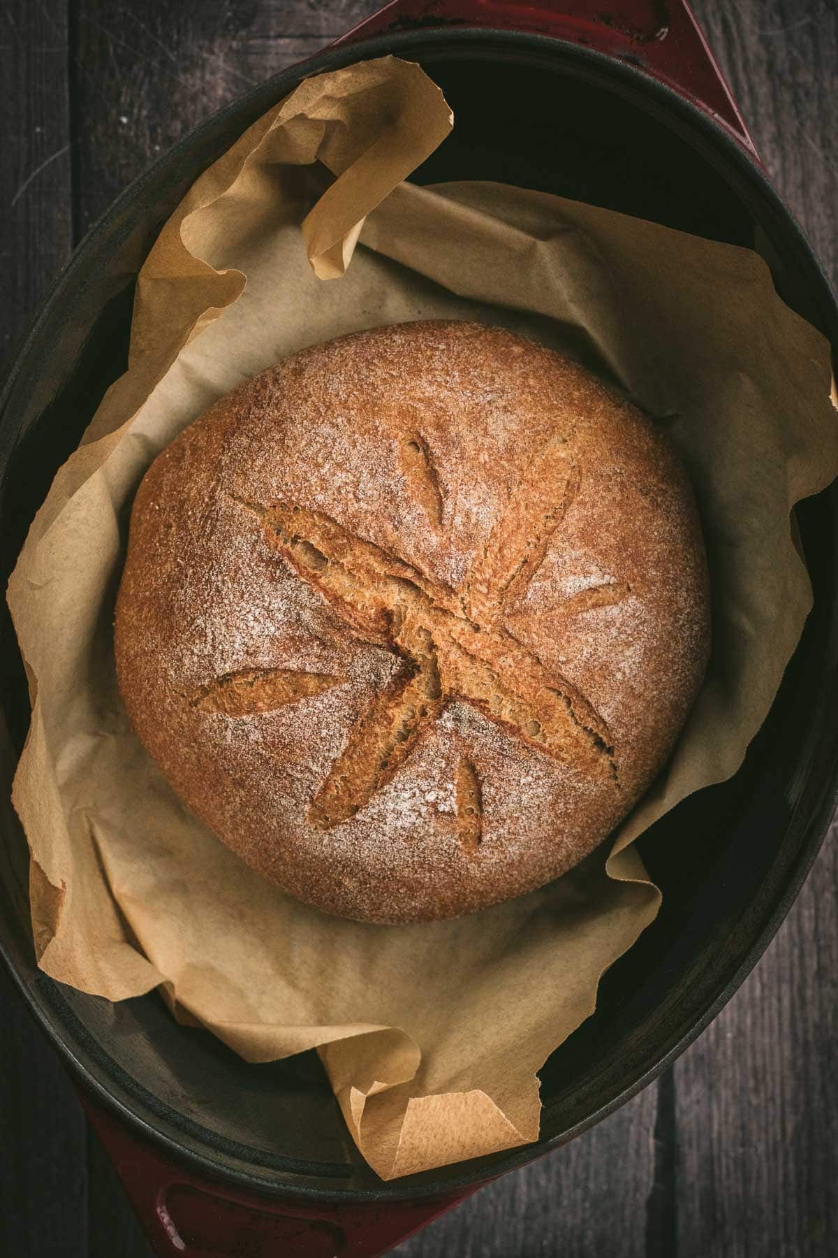A baked loaf of dark brown bread on parchment paper in a Dutch oven.