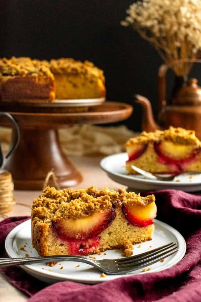 Two slices of plum cake on plates next to more cake on a cake stand.