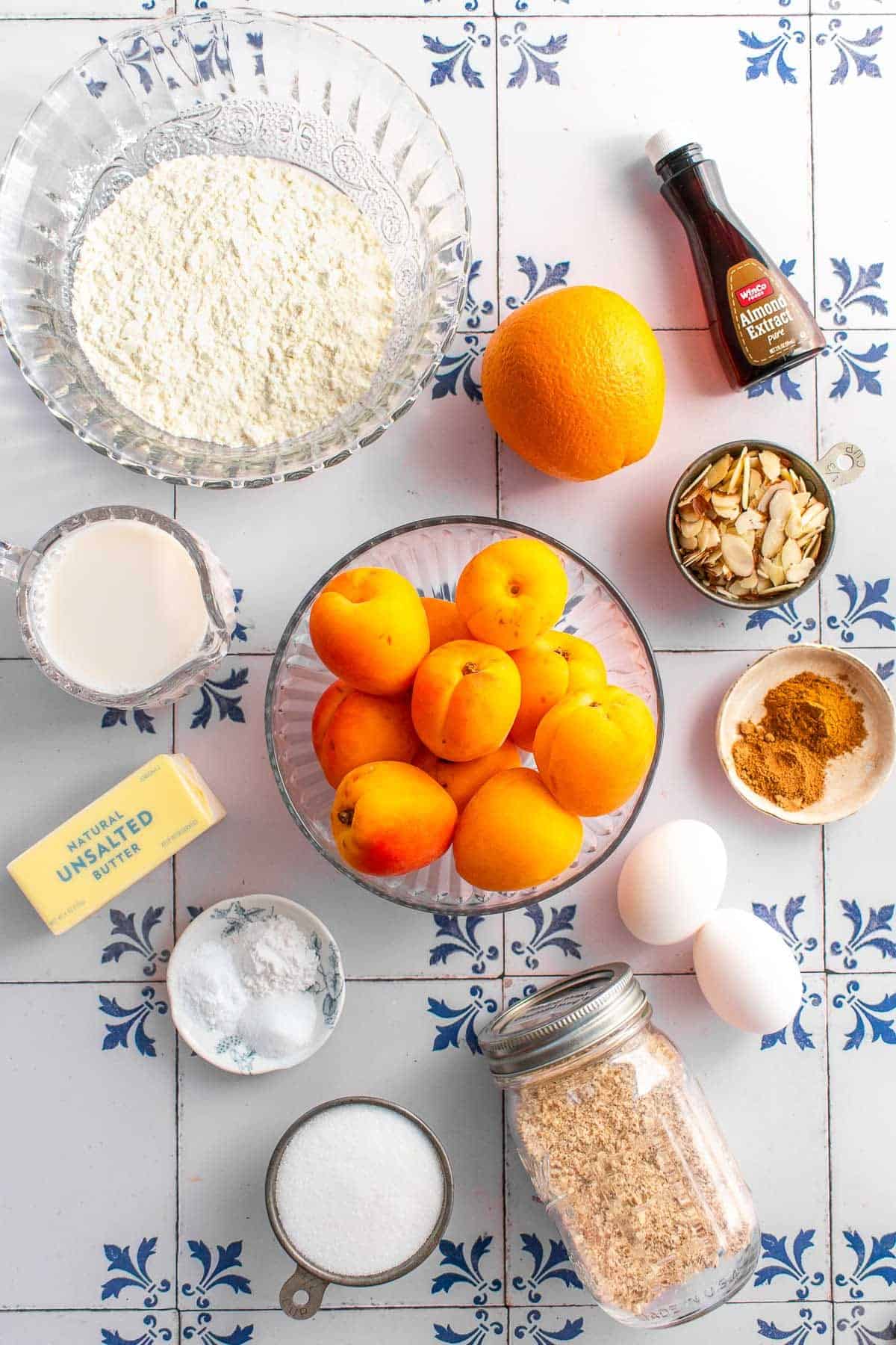 A bowl of apricots next to bowls of flour, sliced almonds, spices, sugar, almond flour, baking powder, salt, and an orange.