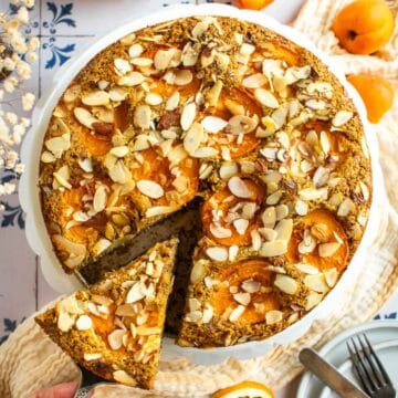 A hand lifting up a slices of almond apricot cake from a cake stand.