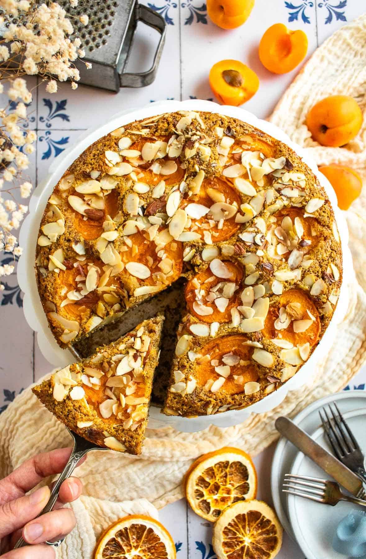 A hand lifting up a slices of almond apricot cake from a cake stand.