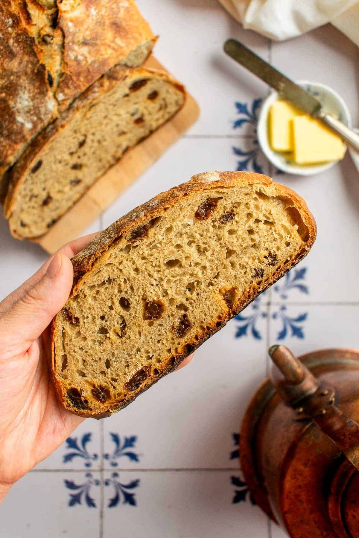 A hand holding a slice of cinnamon raisin bread by a tea kettle.
