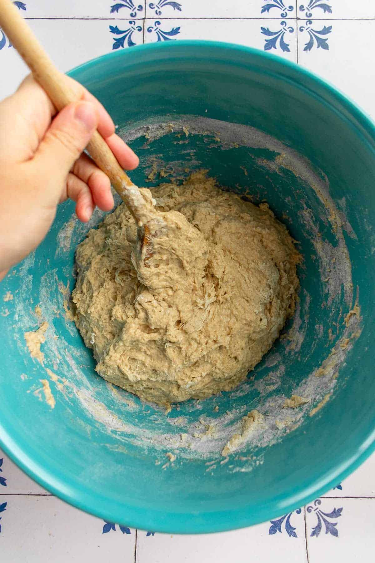 A hand mixing bread dough with a wooden spoon.