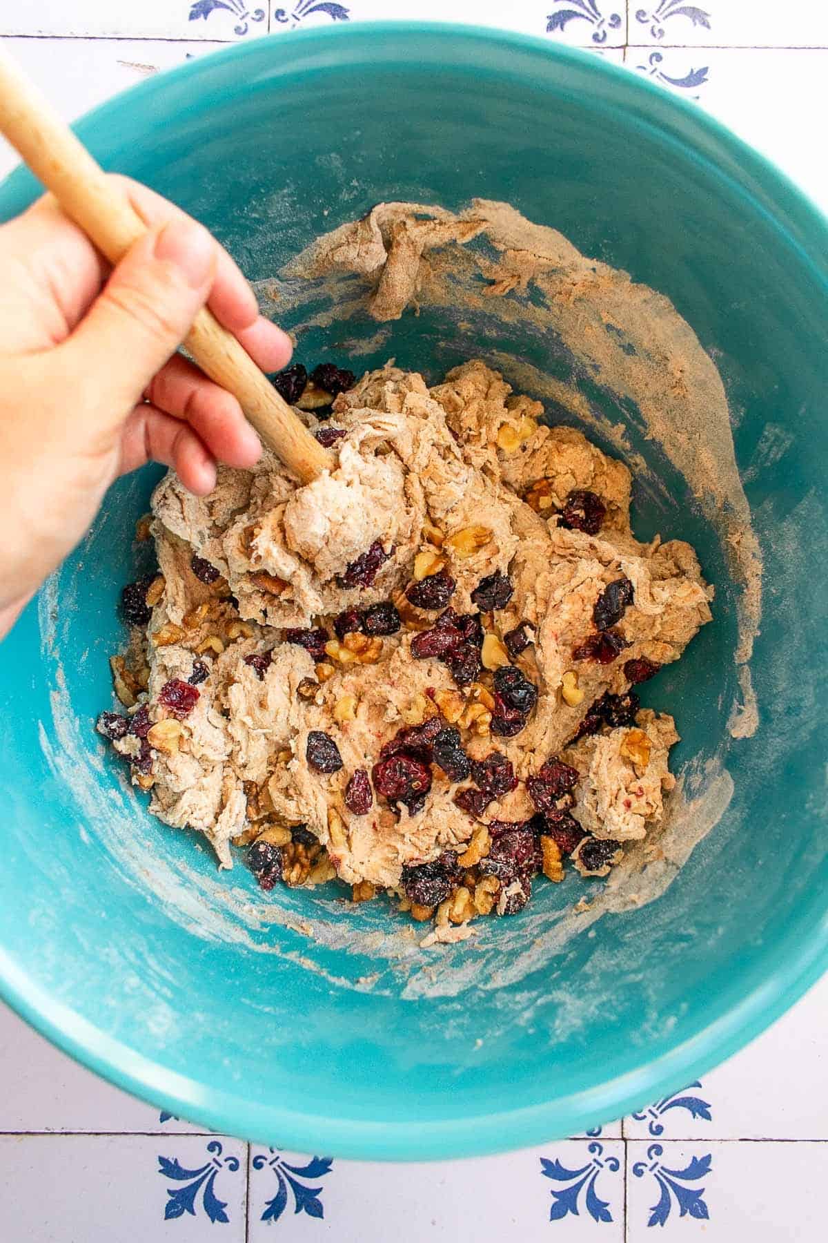 A hand stirring cranberries and walnuts into bread dough with a wooden spoon.
