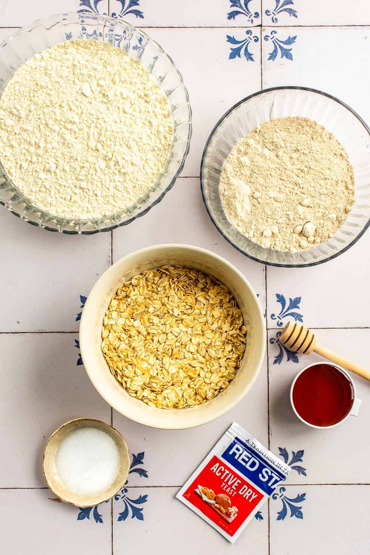 Bowls of white flour, wheat flour, salt, honey, and rolled oats on a table. 