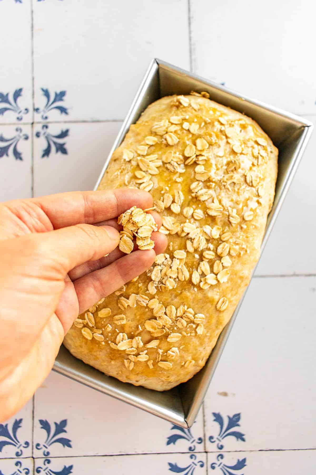 A hand sprinkling rolled oats on top of an unbaked loaf of bread.
