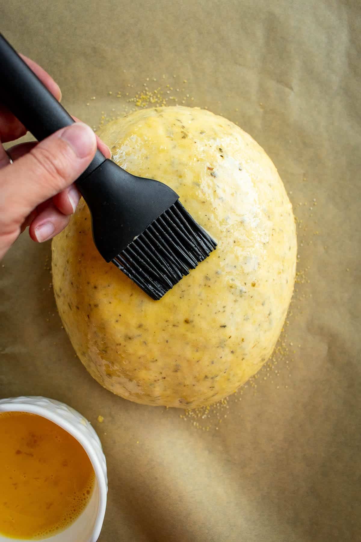 A hand brushing whisked eggs on a loaf of unbaked bread.