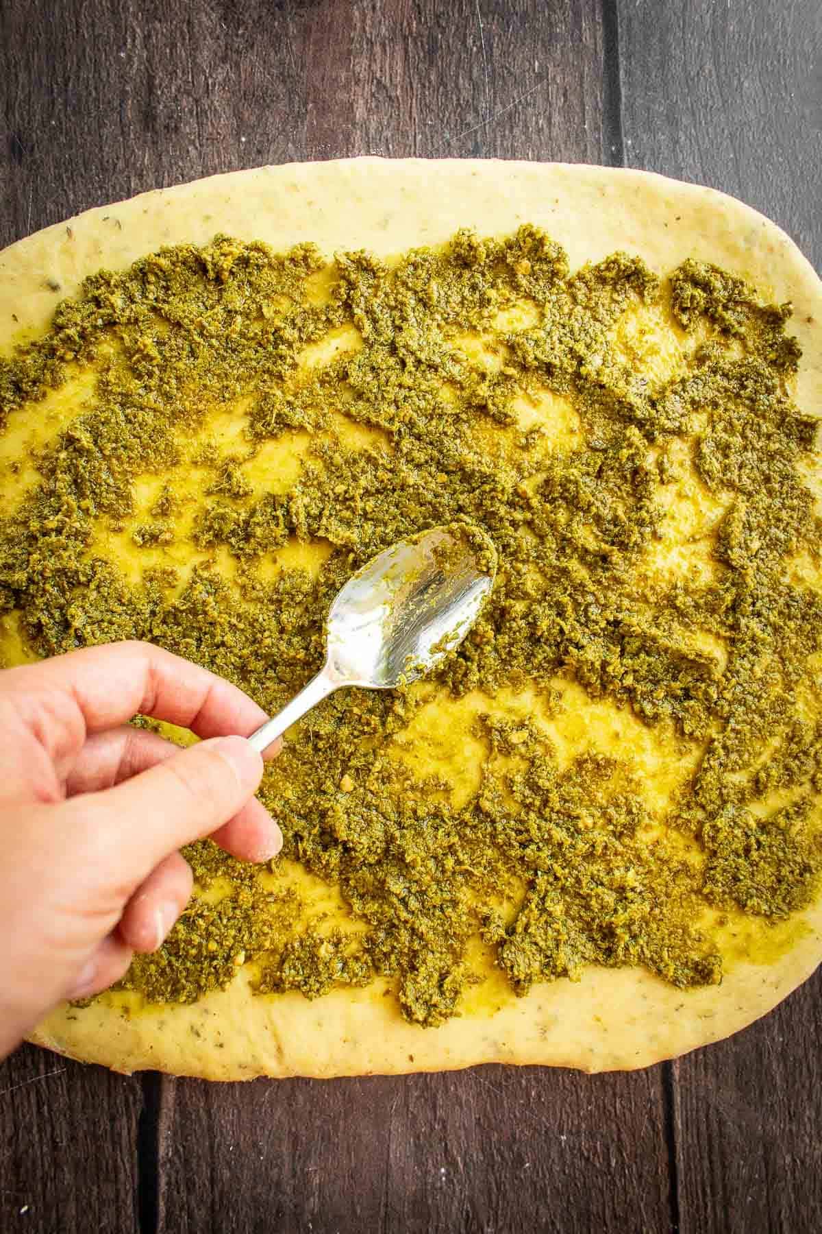 A hand spooning green pesto onto rolled-out bread dough.