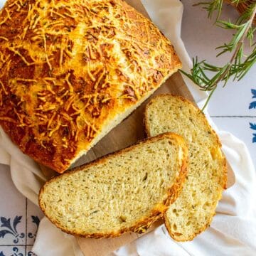 Two slices of bread next to a loaf covered in parmesan and rosemary.