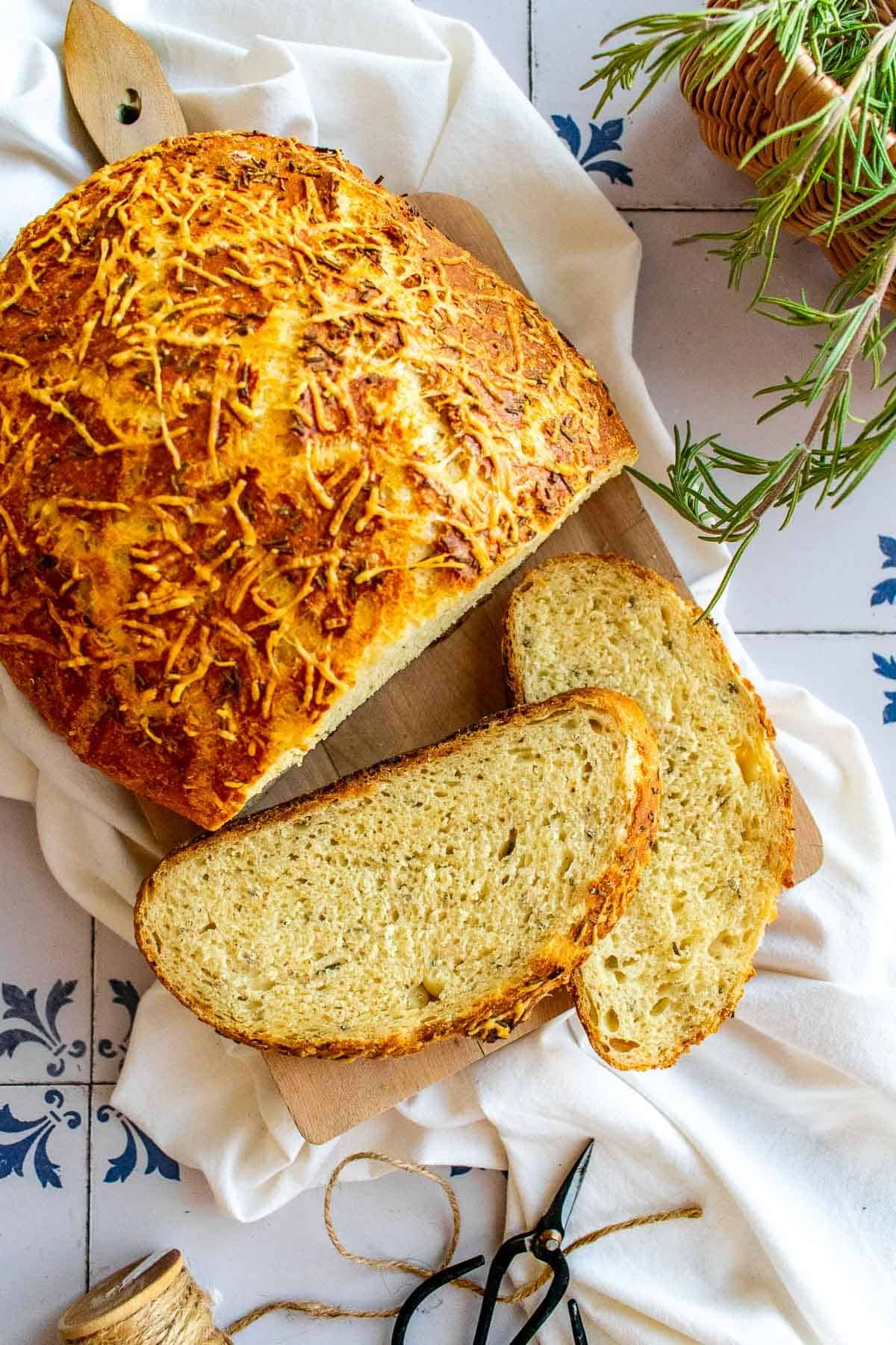 Two slices of bread next to a loaf covered in parmesan and rosemary.
