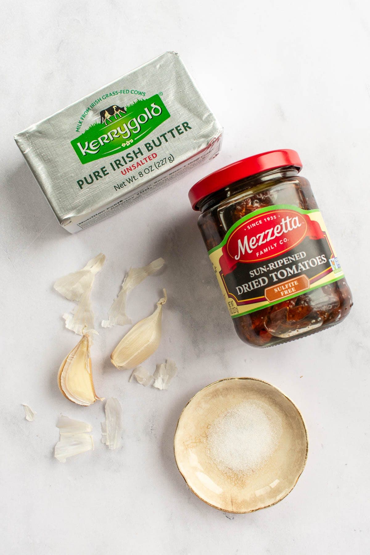 A jar of sun dried tomatoes next to garlic cloves, salt, and a dish of butter.
