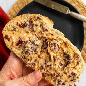 A hand holding a slice of bread covered in butter and sun dried tomatoes.