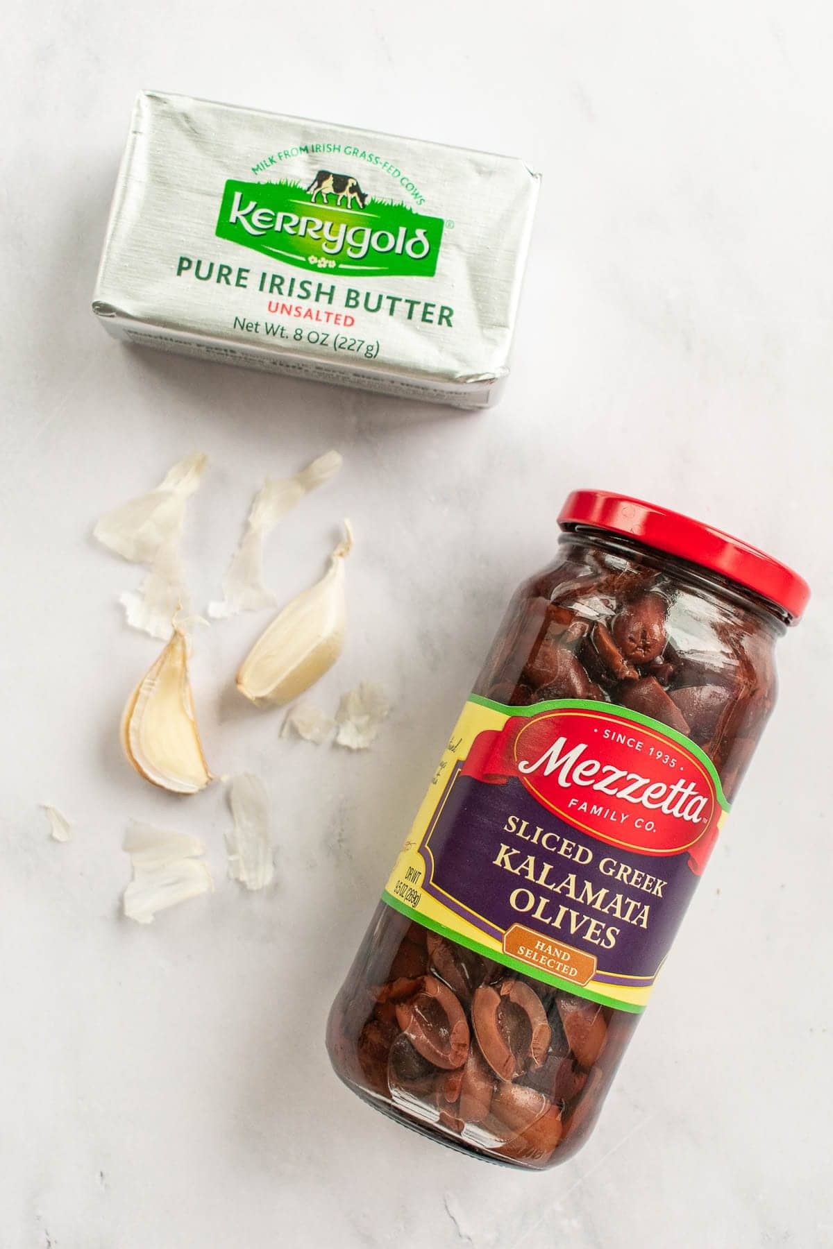 A jar of kalamata olives next to garlic cloves and butter on a table.