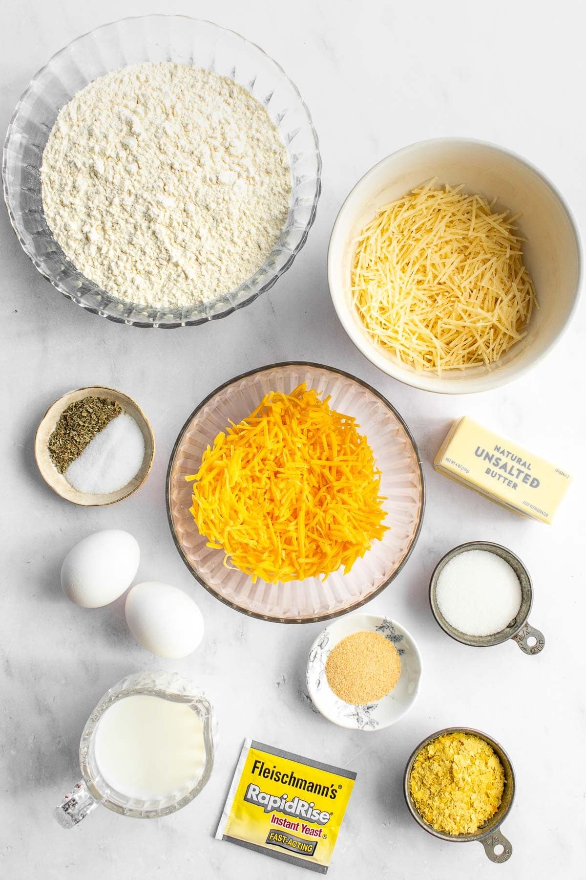 Bowls of cheese, flour, sugar, nutritional yeast, salt, and herbs next to eggs and butter on a table.