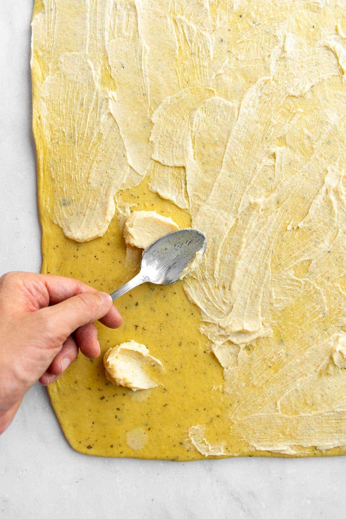 A hand spreading garlic butter onto rolled-out bread dough with a spoon.